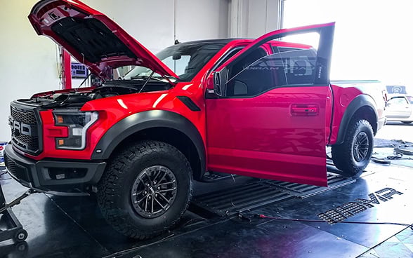 Red and black custom Ford Raptor with open hood and door at auto repair shop.